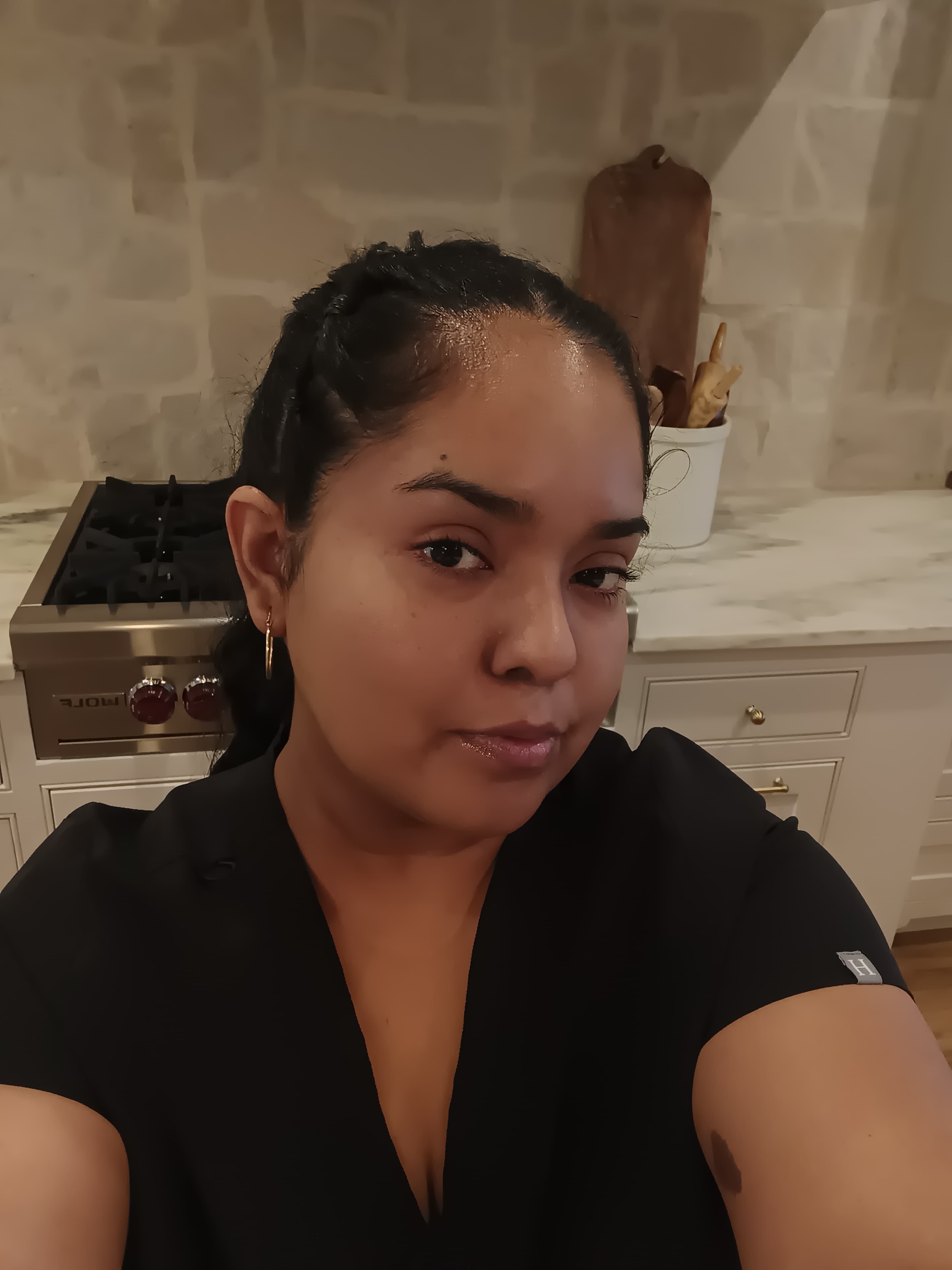Woman with dark braided hair wearing black scrubs in a kitchen with a stone backsplash.